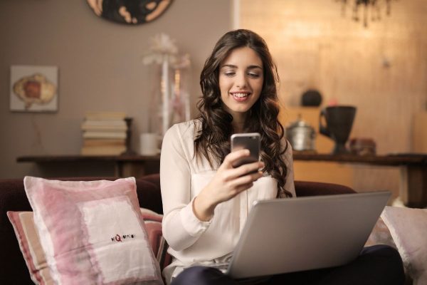 Woman with laptop on couch smiling at smartphone